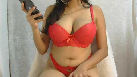Video of a woman with long dark hair, fair skin, wearing red lace lingerie, including a push-up bra and matching panties, posing in a plush chair.