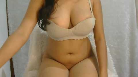 Video of a curvy woman with light skin, long dark hair, wearing beige lingerie, sitting on a white chair with a fluffy blanket draped behind.