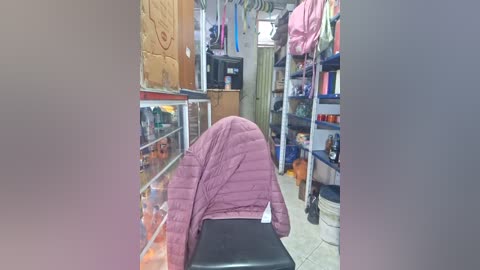 Video of a narrow, cluttered storage room with a person wearing a pink puffy jacket and black pants, bent over a black box, surrounded by shelves and hanging clothes.