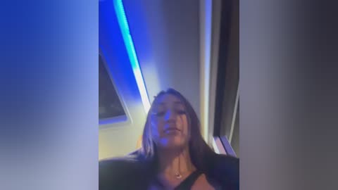 Video of a woman with long, dark hair and fair skin, wearing a black top, standing in a dimly lit room with blue neon lighting and grey curtains.