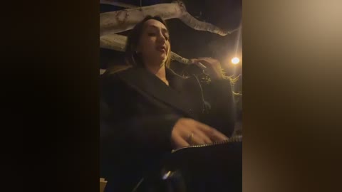 Video of a woman with dark hair, dressed in a dark coat, holding a flashlight, illuminated by a dimly lit, rustic cabin interior with wooden beams and a glowing lamp.