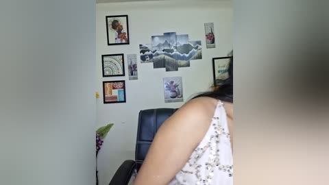 A video of a woman with light skin and dark hair, wearing a sleeveless white dress, standing in front of a wall adorned with framed art, including a large cloud mural and various smaller pieces.