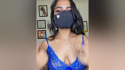 Video of a young woman with medium brown skin, wearing a blue lace bra, black face mask, and a silver necklace. She stands indoors with framed artwork behind her.