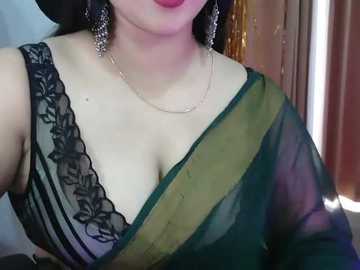 Video of a woman with light skin, wearing a green sari with black lace trim, gold chain necklace, and dangling earrings, indoors, blurred background.