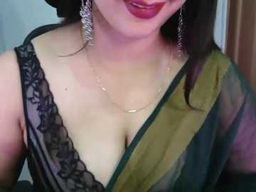 Video of a South Asian woman with light skin, wearing a black lace bra, gold necklace, and green sari, with red lipstick, set against a blurred indoor background.