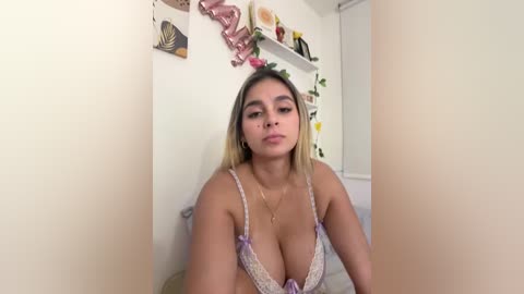 A video of a young woman with light skin and long blonde hair, wearing a lacy lavender bra, in a cozy bedroom with floral decorations.