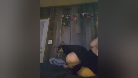 A video shows a bald, overweight man in black clothes, wearing a dog collar and leash, kneeling beside a Doberman Pinscher on a bed. The background features a wooden wall and festive garlands.