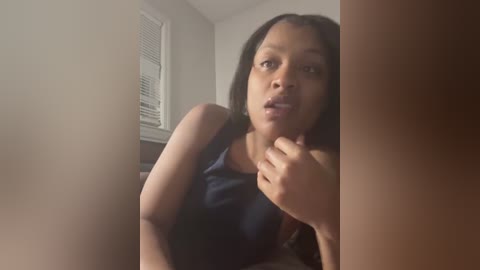 Video of a young woman with medium brown skin and long black hair, wearing a dark blue sleeveless top, sitting indoors near a window with blinds. She has a contemplative expression, with her hand to her mouth.
