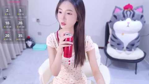 A video of an East Asian woman with long brown hair, fair skin, and small breasts, wearing a floral dress, drinking from a red cup. Background includes a white chair and gray curtains.