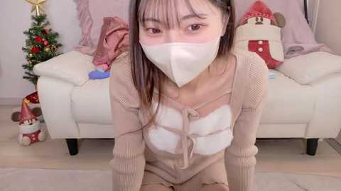 Video of an East Asian woman with long black hair, wearing a beige knit sweater with white reindeer design, a face mask, and kneeling on a beige carpet in a festive, cozy living room with a Christmas tree, decorations, and plush toys.