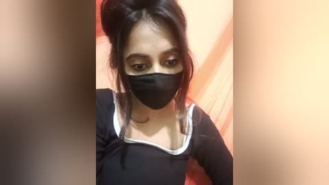 Media: Video of a young woman with light brown skin, black hair in a bun, wearing a black mask and a black long-sleeve shirt with white trim, standing indoors against a beige wall.