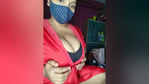 Media: Video of a young woman with medium brown skin and medium-sized breasts wearing a red satin robe, black bra, blue polka-dot mask, and blue polka-dot headband, sitting in a dimly-lit room.