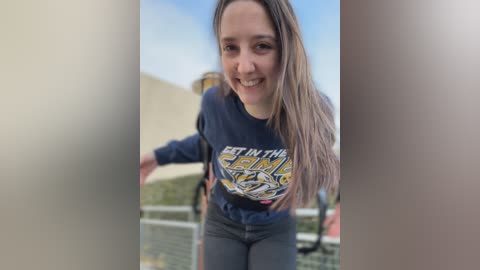 Video of a smiling young woman with long brown hair, wearing a navy blue \"Get in the Game\" t-shirt, standing on a balcony with a clear blue sky background.
