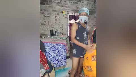 Video of a person in a blue surgical mask, gray tank top, and orange pants, standing in a cluttered, brick-walled room with colorful blankets and a cluttered table.
