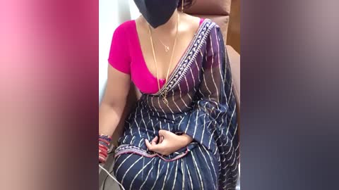 Media: Video of an Indian woman wearing a bright pink short-sleeve blouse, black mask, and traditional blue saree with white stripes. She sits in a car, holding a smartphone, with blurred background.