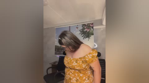 Video of a woman with long brown hair, wearing a yellow floral off-shoulder dress, standing in a dimly lit room with a window displaying a blooming plant.