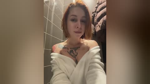 Media: A video of a young woman with short, auburn hair, wearing a white towel, revealing a tattoo on her chest, standing in a tiled bathroom with a brown towel hanging.