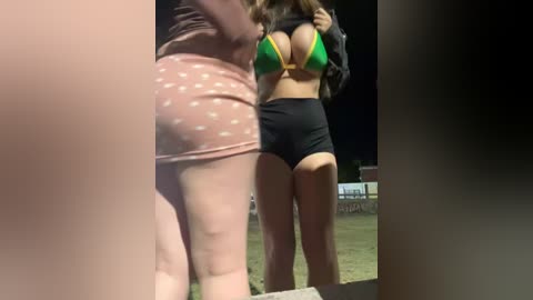 Video of two women at a nighttime sports event; one in a green bikini top and black shorts, the other in polka-dot shorts, with a blurred stadium background.