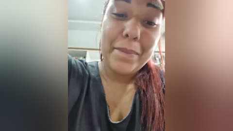 Video of a young Asian woman with fair skin, long red hair, and dark eyes, smiling, wearing a dark T-shirt. Background shows blurred, indoor setting with neutral colors.