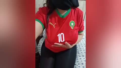 A video of a woman wearing a red football jersey with a green crest and number 10, black leggings, and a black mask, sitting on a bed with polka dot sheets.