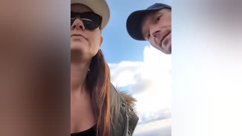 Video of a woman in sunglasses, beige hat, and green jacket, and a man in a cap, looking down at a partially blurred person in the foreground against a bright, clear sky.