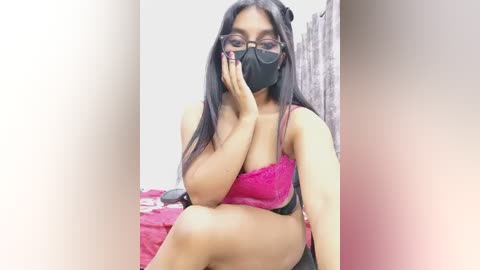 Video of a South Asian woman with long black hair, wearing a pink lace bra and black mask, sitting on a bed with a pink sheet, in a dimly lit room.