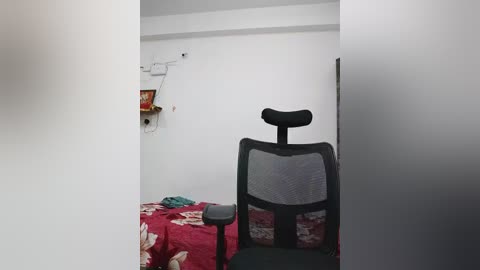 Video of a sparsely furnished room with a white wall, black office chair, red floral tablecloth, and a mounted shelf with a phone and books.