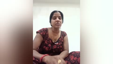 A video of a middle-aged woman with medium brown skin, sitting indoors, wearing a floral-patterned red dress. She has short, dark hair, and her eyes are closed, appearing contemplative.