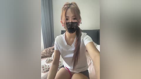 Video of an Asian woman with light brown hair in a ponytail, wearing a white T-shirt and pink panties, seated on a bed with a black headboard, in a softly lit bedroom.