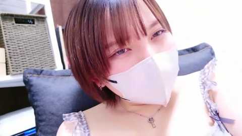 A video of a young Asian woman with straight brown hair and bangs, wearing a white surgical mask, a delicate necklace, and a lace-trimmed white top. She sits on a dark blue cushioned chair in a modern room with a woven basket and white walls.