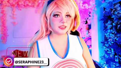 Video of a young woman with long, pastel pink hair, wearing a white crop top with blue trim and a large pink heart graphic, set against a vibrant, multi-colored background.