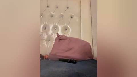 Video of a beige upholstered bed with a tufted, padded headboard. A large, pink pillow and a black hairbrush are placed on the bed. The room's walls are painted in a soft, light color.