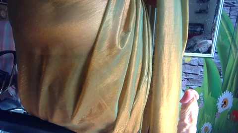 Close-up video of a person wearing a shiny, golden-yellow shirt, with a brick wall and green floral-patterned fabric in the background.