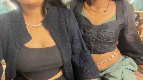 Video of two women in casual attire: One in a navy blue cardigan over a dark top, and the other in a dark blouse with a beaded necklace. Background is blurred.