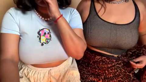Video of two women sitting close, one in a light blue crop top with a colorful cartoon face and beige pants, the other in a gray tank top with a leopard print skirt.