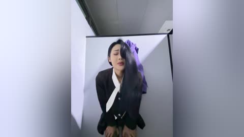 Video of a young Asian woman with long black hair, wearing a black suit and white tie, kneeling on a white tiled floor, eyes closed, in a dimly lit, stark white room with a large mirror.
