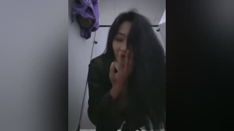 Video of a young Asian woman with long black hair, wearing a black jacket, covering her face with her hands, in a dimly lit bathroom with purple clothes hanging on the wall.