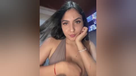 Video of a young, light-skinned South Asian woman with long black hair, wearing a brown halter top, resting her chin on her hand, indoors with a blurred background.