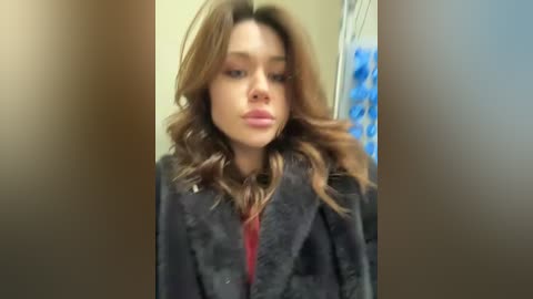 Video of a young woman with light brown, wavy hair, wearing a dark coat over a red top, standing indoors with blurred background.