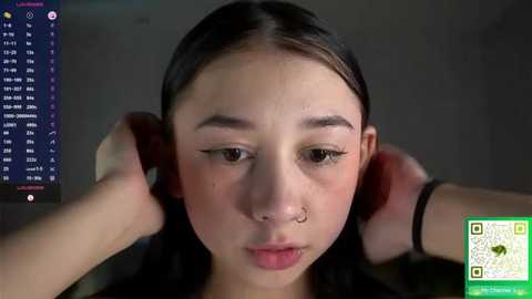 Video of an Asian woman with fair skin, long dark hair, and a septum piercing, wearing a black top, adjusting headphones while looking at a computer screen.