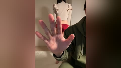Video of a woman wearing a white blouse and red shorts standing in a dimly lit room. A man in a green hoodie and bare feet is visible in the foreground, with his hand blocking the view.