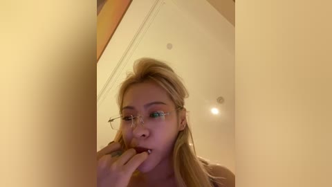 Media: A video of a blonde woman with glasses, wearing a sleeveless top, standing in front of a mirror, biting her finger, in a dimly lit room with beige walls.