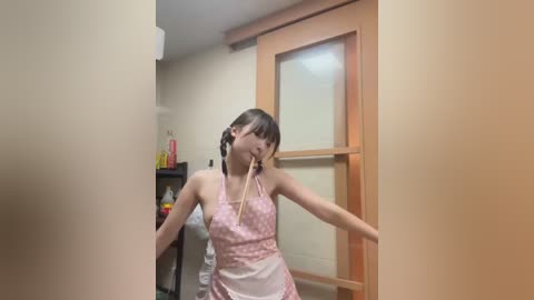 Media: A video of an Asian woman with brown hair in braids, wearing a pink polka-dot apron, holding chopsticks, standing in a kitchen with wooden cabinets and a stove.