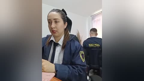 Video of a young woman with fair skin and brown hair tied back, wearing a dark navy blue police uniform and a black tie, seated in a room with another officer in the background.