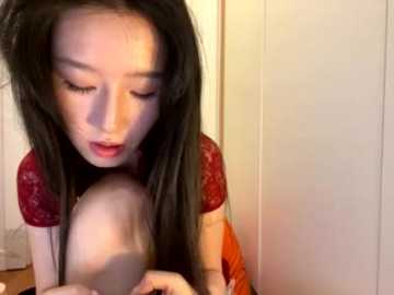A video of an East Asian woman with long black hair, fair skin, and wearing a red lace top, sucking on a large, bare breast, in a dimly lit room with white walls.