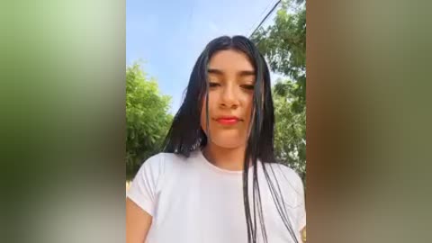 A video of a young woman with long black hair, wearing a white t-shirt, standing outdoors with green foliage and a clear blue sky in the background.