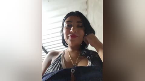Video of a South Asian woman with medium skin tone, black hair, wearing a black halter top, standing in a dimly lit room with a window and concrete wall.