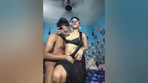 Video of two people in a bedroom. A shirtless man with medium skin tone and short black hair kisses a woman with a medium skin tone, wearing a black bra and black pants.