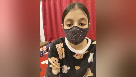 Video of a young girl with light brown skin, dark hair, wearing a black face mask and a pajama top with teddy bears, standing in a room with red curtains and a plush toy on the floor.
