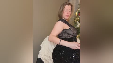 A video of a fair-skinned, plus-sized woman with shoulder-length brown hair, wearing a sheer black lace top and a black skirt with silver sequins, standing near a Christmas tree in a softly lit room.